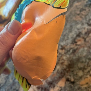 May include: A yellow, blue, and red plastic mask with a torn edge. The mask is shaped like a face with a large nose and a wide mouth.