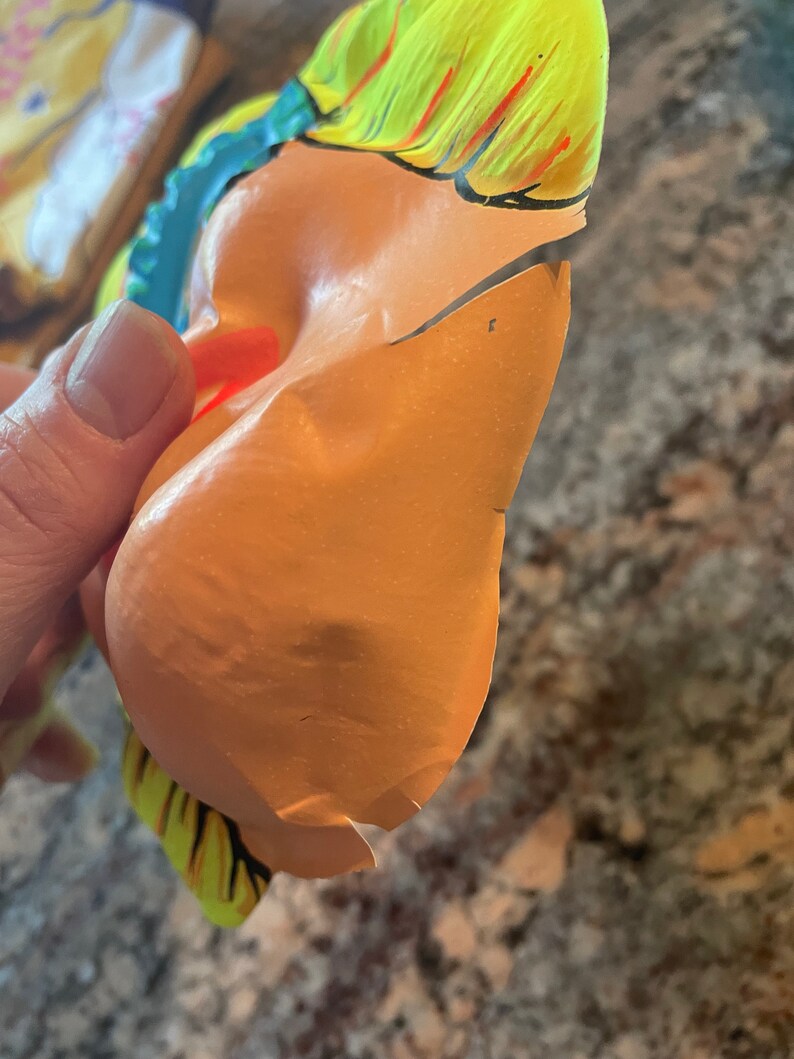 May include: A yellow, blue, and red plastic mask with a torn edge. The mask is shaped like a face with a large nose and a wide mouth.