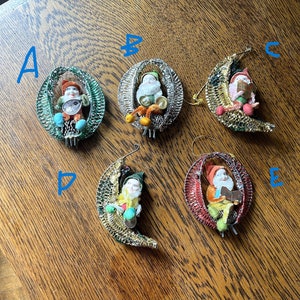 May include: A collection of vintage Christmas ornaments featuring small figures inside decorative wire frames and crescent moons. The ornaments depict elves and Santa Claus-like characters, with colorful clothing and accessories. These ornaments are perfect for holiday decor.