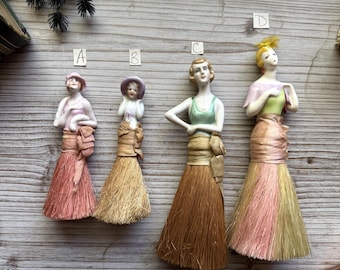 May include: Four vintage porcelain figurines of women, each with a unique hairstyle and attire. The figures have brush-like skirts in shades of pink, gold, and brown. The figurines are displayed on a wooden surface.