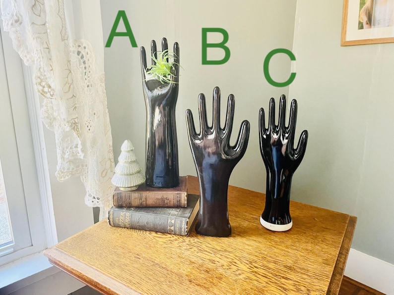 May include: Three black ceramic hand-shaped ring holders. The middle holder is the largest and the left holder has a small green plant inside.