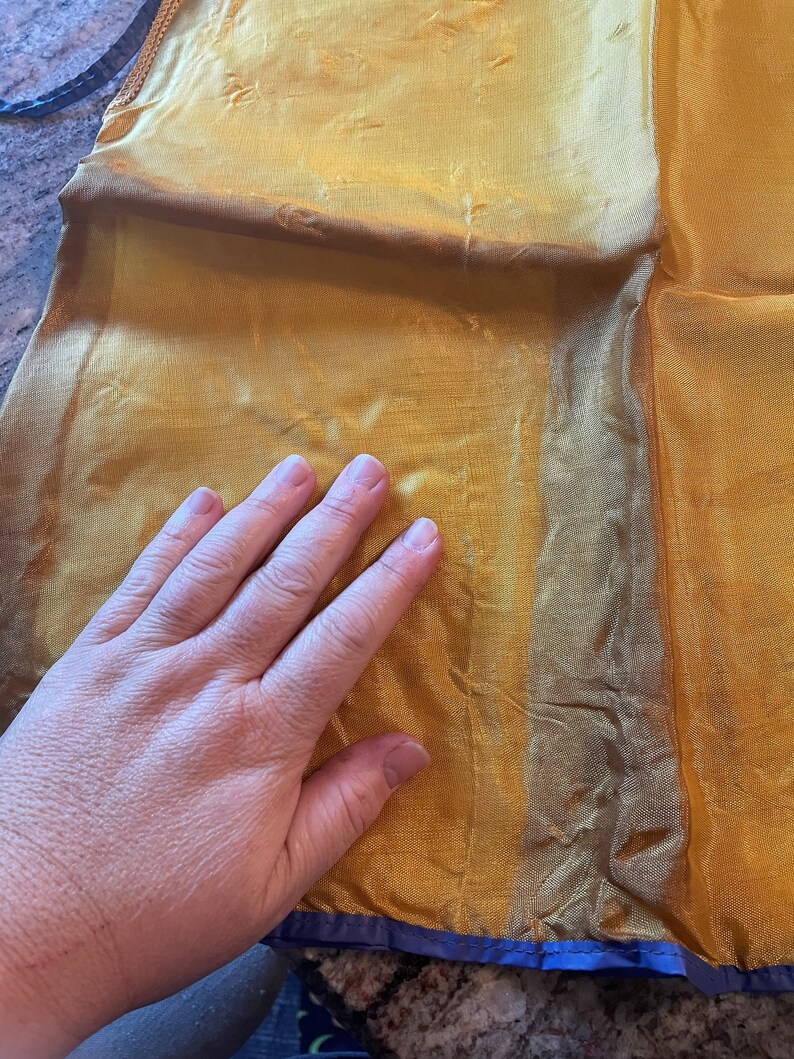 May include: A gold sheer fabric with a blue trim. The fabric is folded in half and a hand is resting on top of it.