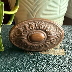 May include: An antique bronze oval door knob with ornate floral detailing. The knob features a central oval and is set against a textured green surface. The knob's aged appearance suggests it is vintage.