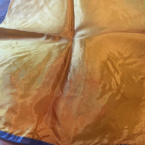 May include: A gold silk scarf with a navy blue trim. The scarf is folded in half and is being held by a hand.
