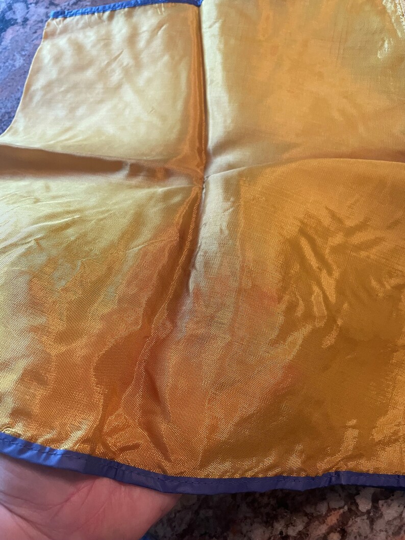 May include: A gold silk scarf with a navy blue trim. The scarf is folded in half and is being held by a hand.