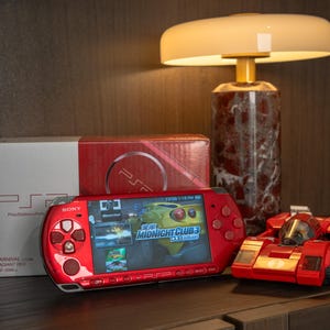 Puede incluir: Una consola de juegos portátil Sony PSP roja con un juego que se muestra en la pantalla, una caja roja y blanca y un coche de carreras de juguete rojo. Una lámpara con base de mármol y pantalla blanca en el fondo.