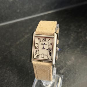May include: A rectangular Seiko watch with a cream face, black Roman numerals, and a date display. The watch has a silver case and a light tan suede strap. The watch is displayed on a clear stand.