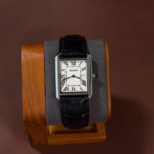 Pode incluir: Um relógio de pulso retangular Seiko com caixa prateada, mostrador branco com algarismos romanos pretos e pulseira de couro preto texturizado. O relógio inclui uma janela de data e uma coroa azul na lateral.