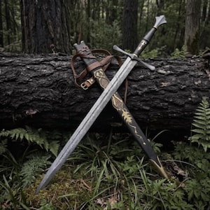 May include: A steel longsword with a crossguard and a dark handle, featuring a pointed blade and a matching black scabbard adorned with gold-colored filigree patterns and brown leather straps with metal buckles for secure carrying and storage.