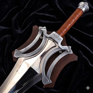 May include: A silver-toned metal sword featuring a complex, multi-layered guard with curved accents and a brown leather-wrapped handle. The crossguard displays engraved text. The sword is mounted on a dark brown wooden plaque with a curved shape.
