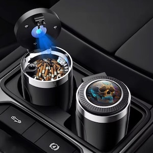 Puede incluir: Dos ceniceros negros para coche. Uno está abierto, revelando colillas de cigarrillos y una luz azul. El otro está cerrado, con un diseño de calavera. Ambos tienen un borde plateado y están en una consola de coche.