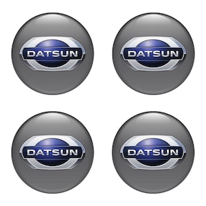 May include: Four grey hubcaps featuring the Datsun logo. Each circular hubcap has a silver border and a blue and white Datsun emblem in the centre. The hubcaps are arranged in a 2x2 grid.
