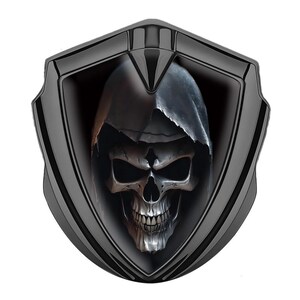 May include: A black shield-shaped emblem featuring a detailed skull wearing a hooded cloak. The skull has visible teeth and eye sockets, with a dark background and a glossy black frame. The design is a gothic or biker-themed emblem.
