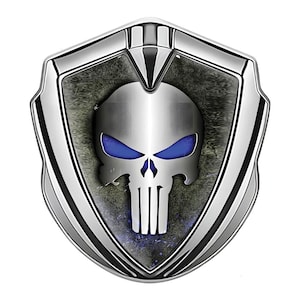 May include: A silver shield-shaped emblem featuring a skull with blue eyes. The skull is centered on a textured gray background, framed by a shiny silver border. The design has a metallic, three-dimensional appearance.