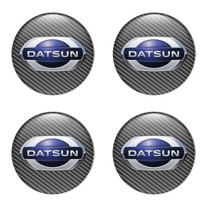 May include: Four Datsun logo emblems. Each emblem features the Datsun logo in a blue and silver oval shape, set against a carbon fiber background. The emblems are circular and have a glossy finish.