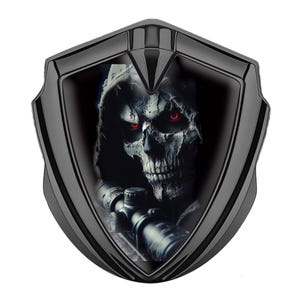 May include: Black shield-shaped emblem featuring a grim reaper skull with red eyes and a sniper scope. The skull is detailed with a hood and a skeletal face. The emblem has a glossy finish.