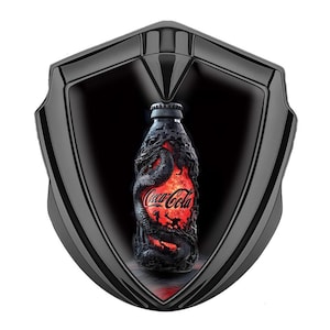 May include: A black shield-shaped display case frames a Coca-Cola bottle. The bottle is intricately detailed with a dragon design, and the Coca-Cola logo is visible. The bottle has a red and orange glow, and the background is black.