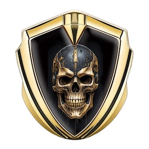 May include: A gold and black shield-shaped emblem featuring a detailed skull design. The skull has a metallic appearance with gold and dark gray accents, set against a black background. The outer frame is a polished gold.