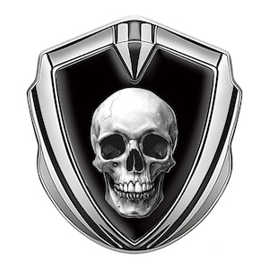 May include: A detailed illustration of a skull within a shield-shaped emblem. The skull is rendered in grayscale with intricate shading, set against a black background. The shield has a polished silver border, creating a striking contrast.