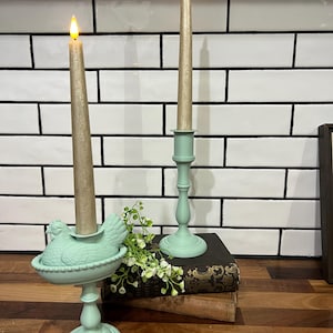 May include: Two decorative candle holders with lit candles. One holder is a light green color and shaped like a hen. The other is a traditional candlestick design. The candles are a light gold color.