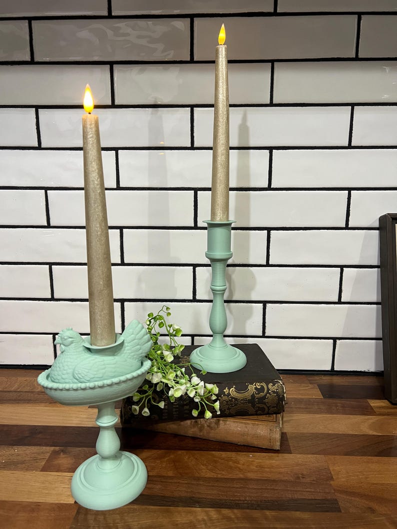 May include: Two decorative candle holders with lit candles. One holder is a light green color and shaped like a hen. The other is a traditional candlestick design. The candles are a light gold color.