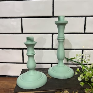 May include: Two mint green candle holders with a classic design. The candle holders are made of a solid material and have a threaded top for securing candles. They are displayed on a dark surface against a white tile background.
