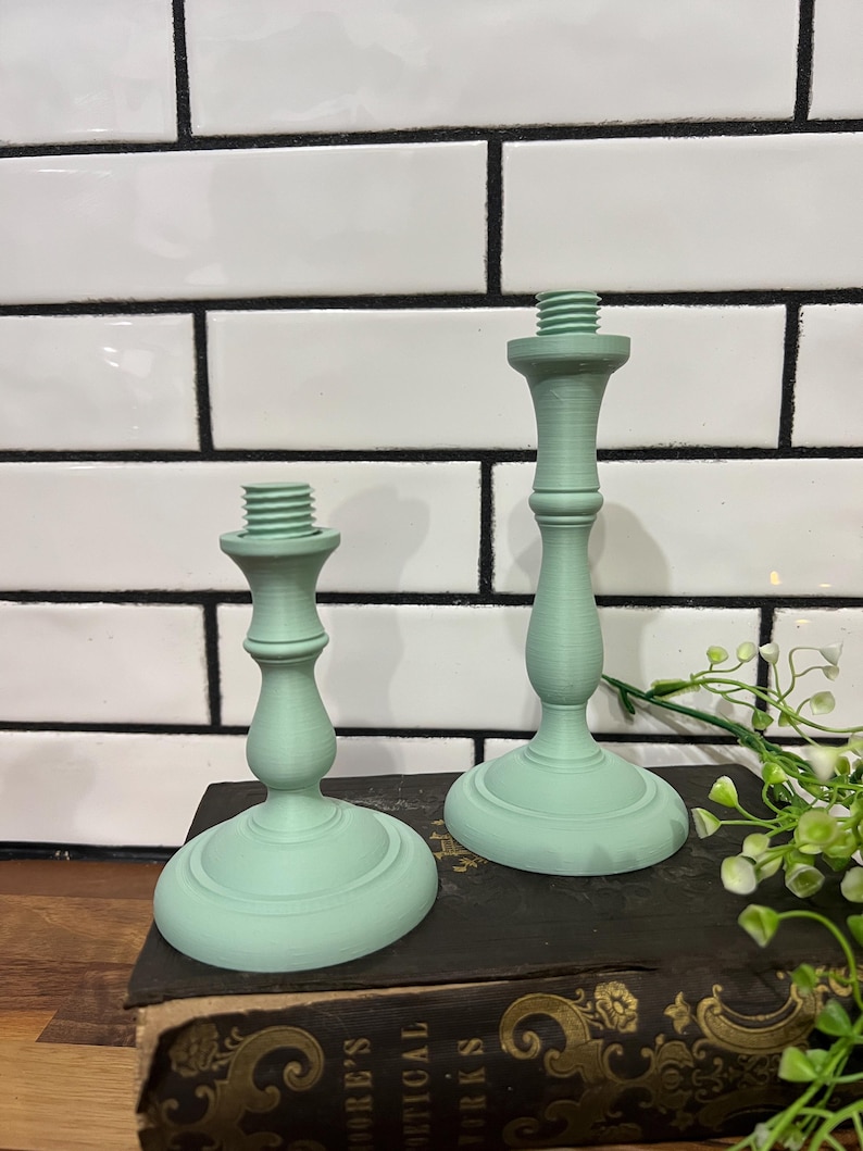 May include: Two mint green candle holders with a classic design. The candle holders are made of a solid material and have a threaded top for securing candles. They are displayed on a dark surface against a white tile background.