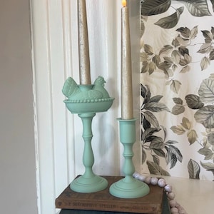 May include: Two decorative candle holders in a soft green hue. One features a hen and nest design, the other a simple cup shape. Both hold tall, gold-colored candles. The items sit atop vintage books, creating a charming, rustic display.