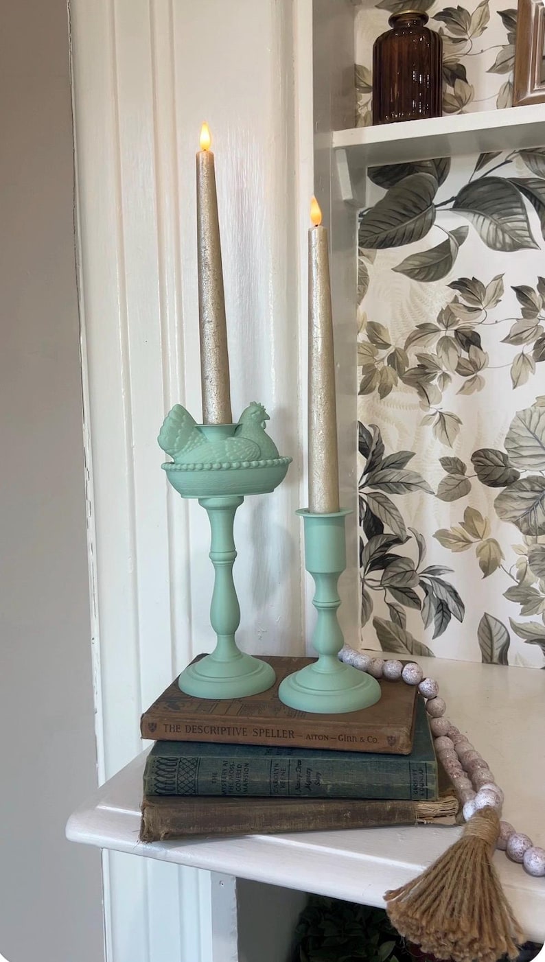 May include: Two decorative candle holders in a soft green hue. One features a hen and nest design, the other a simple cup shape. Both hold tall, gold-colored candles. The items sit atop vintage books, creating a charming, rustic display.