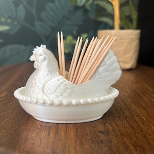May include: A white ceramic chicken-shaped toothpick holder. The holder is in the shape of a hen, with a detailed textured design. The holder is filled with wooden toothpicks. The holder is sitting on a wooden surface.