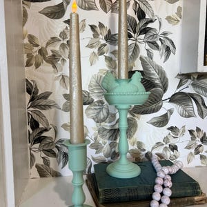 May include: Two light green candle holders with gold candles. One holder has a chicken-shaped design. The candles are lit. A string of white and pink beads and a stack of books are in the foreground. The background has a floral wallpaper.