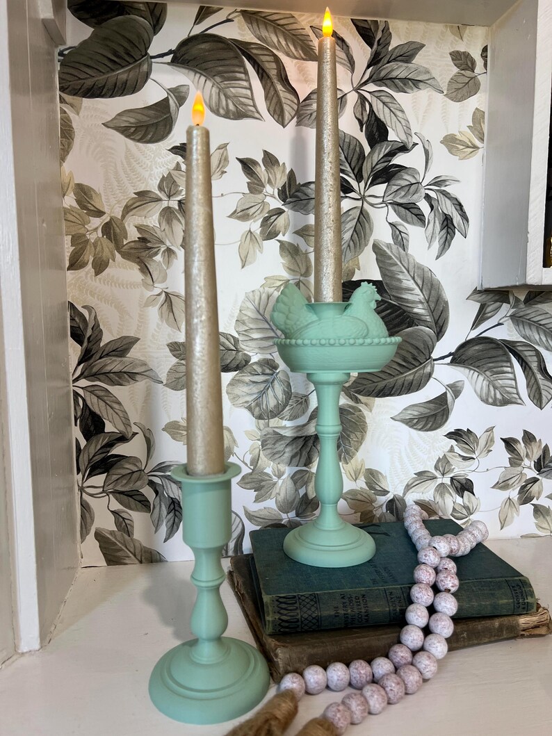 May include: Two light green candle holders with gold candles. One holder has a chicken-shaped design. The candles are lit. A string of white and pink beads and a stack of books are in the foreground. The background has a floral wallpaper.