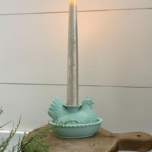 May include: A silver taper candle with a flickering flame sits in a light blue ceramic hen-shaped candle holder. The candle holder is on a wooden surface, with a white wall in the background.