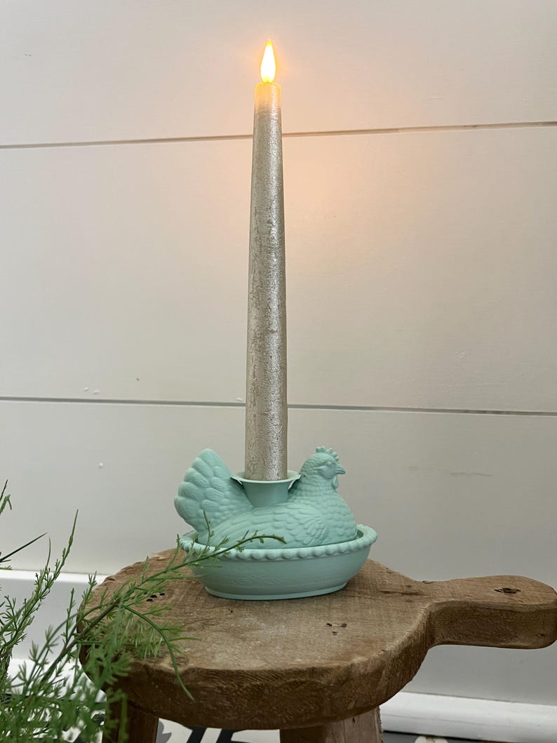 May include: A silver taper candle with a flickering flame sits in a light blue ceramic hen-shaped candle holder. The candle holder is on a wooden surface, with a white wall in the background.