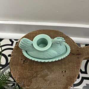 May include: A light teal chicken-shaped butter dish with a round opening on top. The dish sits on a wooden cutting board with a decorative edge. The background includes a white surface and a black and white patterned tile.