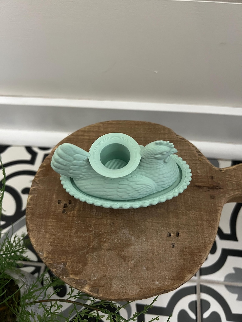May include: A light teal chicken-shaped butter dish with a round opening on top. The dish sits on a wooden cutting board with a decorative edge. The background includes a white surface and a black and white patterned tile.