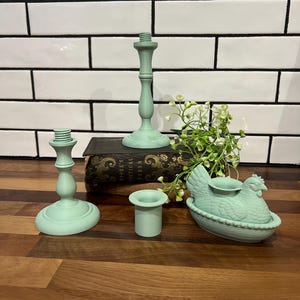 May include: A collection of mint green candle holders and a hen-shaped candle holder. The set includes two tall candlestick holders, a small cylindrical holder, and a hen-shaped holder with a candle cup. The items are arranged on a wooden surface.
