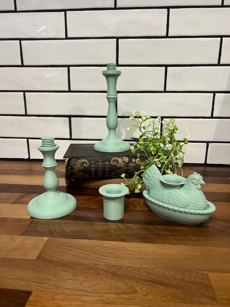 May include: A collection of mint green candle holders and a hen-shaped candle holder. The set includes two tall candlestick holders, a small cylindrical holder, and a hen-shaped holder with a candle cup. The items are arranged on a wooden surface.