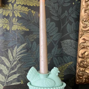 May include: A light green candle holder shaped like a hen sitting on a nest, holding a tall, light pink candle with a flickering flame. The background features a dark wallpaper with a leaf pattern and a gold-framed mirror.