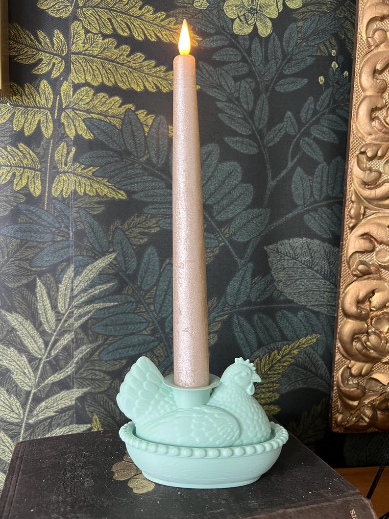 May include: A light green candle holder shaped like a hen sitting on a nest, holding a tall, light pink candle with a flickering flame. The background features a dark wallpaper with a leaf pattern and a gold-framed mirror.