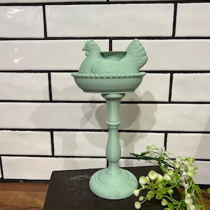 May include: A mint green chicken-shaped candle holder. The hen sits atop a beaded bowl, supported by a pedestal base. The decorative piece is placed on a dark book, with a white-tiled background.