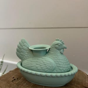 May include: A light teal ceramic hen-shaped candle holder. The hen is sitting in a decorative bowl with a scalloped edge. The candle holder is on a wooden surface.