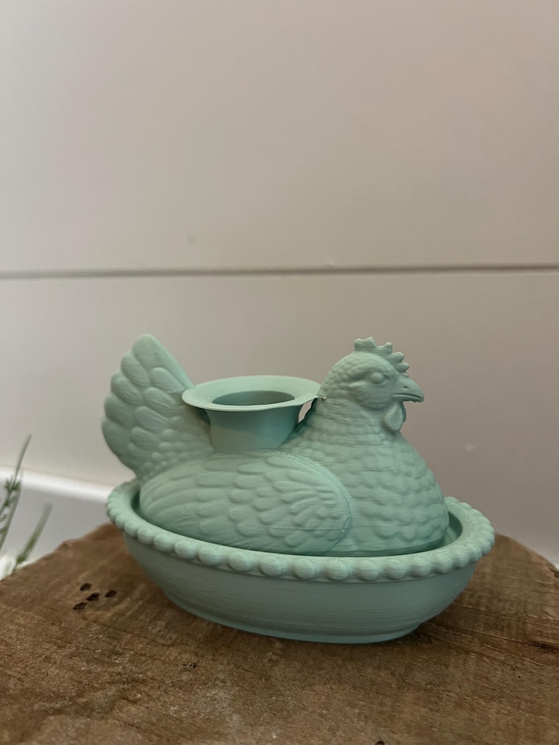 May include: A light teal ceramic hen-shaped candle holder. The hen is sitting in a decorative bowl with a scalloped edge. The candle holder is on a wooden surface.