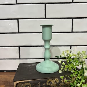 May include: A mint green candlestick holder with a round base and a slender stem. The candlestick is placed on a vintage book with gold lettering. The background features white tiles with black grout and a sprig of greenery.
