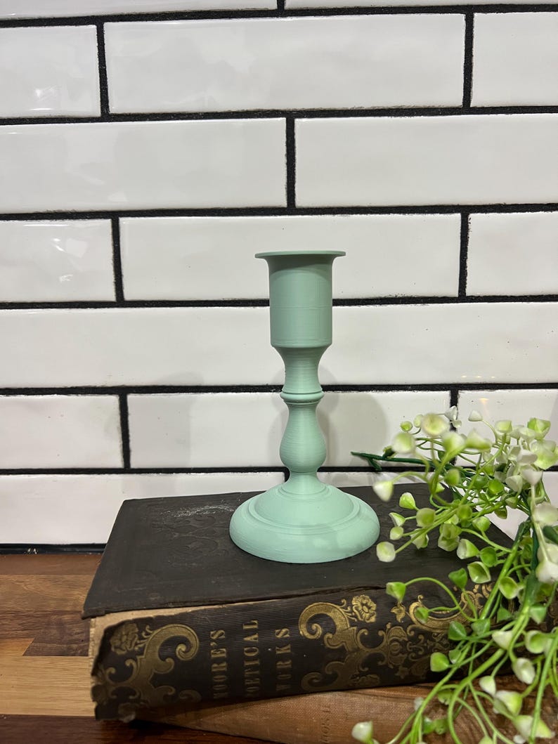 May include: A mint green candlestick holder with a round base and a slender stem. The candlestick is placed on a vintage book with gold lettering. The background features white tiles with black grout and a sprig of greenery.