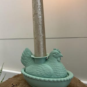 May include: A light teal chicken-shaped candle holder with a silver candle. The candle holder is designed with a detailed hen sitting in a nest. The candle holder is a decorative item.