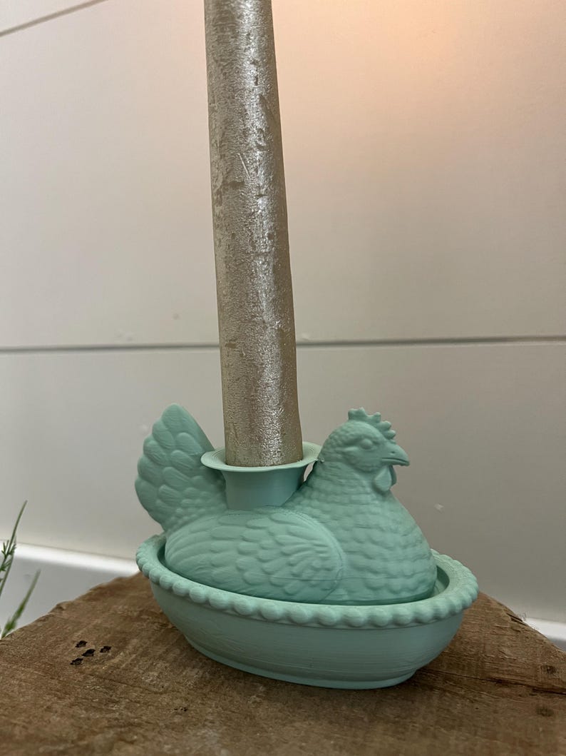 May include: A light teal chicken-shaped candle holder with a silver candle. The candle holder is designed with a detailed hen sitting in a nest. The candle holder is a decorative item.