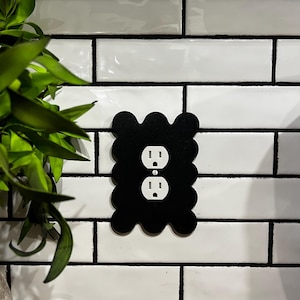 May include: A black, scalloped outlet cover is mounted on a white tile wall. The cover has two openings for electrical outlets. A green plant is visible in the upper left corner of the image.