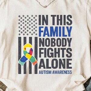 May include: Cream-colored t-shirt with a graphic design promoting autism awareness. The design features the text "IN THIS FAMILY NOBODY FIGHTS ALONE" in blue and black, with a colorful ribbon and the words "AUTISM AWARENESS".