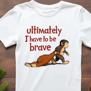 Op de afbeelding: Wit T-shirt met de tekst "Ultimately I have to be brave" in het rood. Het shirt heeft een cartoon illustratie van een aap die een andere aap aan de staart trekt. De illustratie is bruin en bruin.